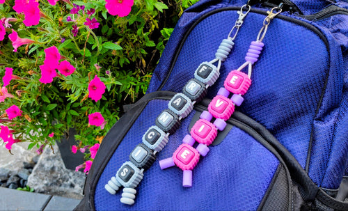 Two personalized letter bead keychains spelling 'FRANCIS' and 'EVIE' hanging from a blue backpack, with bright pink flowers in the background.